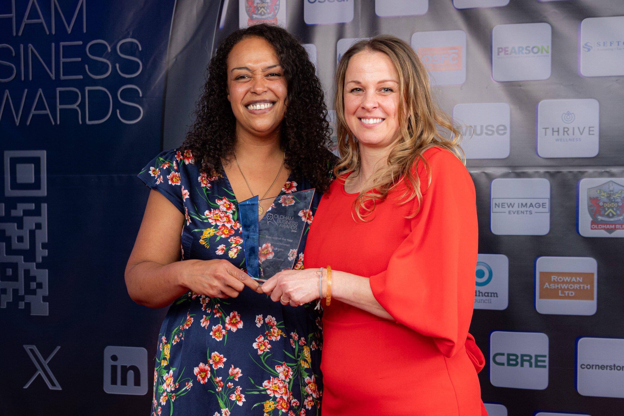 Two women smiling and holding an award together at a business awards event. They stand in front of a backdrop with sponsor logos and the words Business Awards visible. Two women smiling and holding an award together at a business awards event. They stand in front of a backdrop with sponsor logos and the words Business Awards visible.