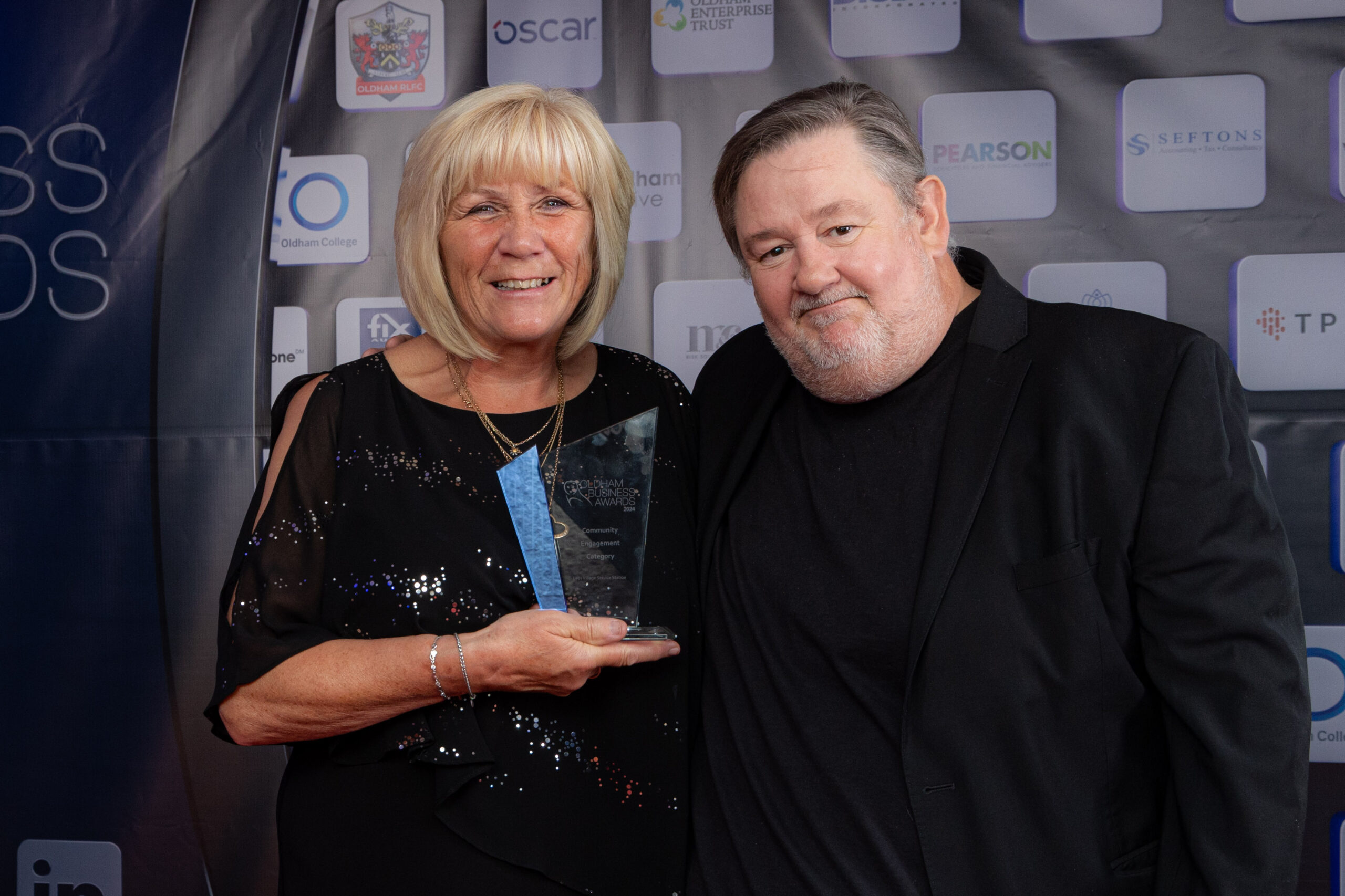 A woman holding a glass award stands next to a man in front of a backdrop with various company logos. Both are smiling and dressed in black attire at what appears to be an awards event.
