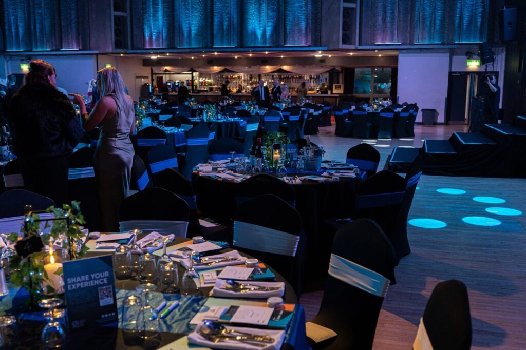 A dimly lit event venue with round tables set for dinner, black chairs with white sashes, and guests mingling near a bar in the background under blue lighting. A dimly lit event venue with round tables set for dinner, black chairs with white sashes, and guests mingling near a bar in the background under blue lighting.