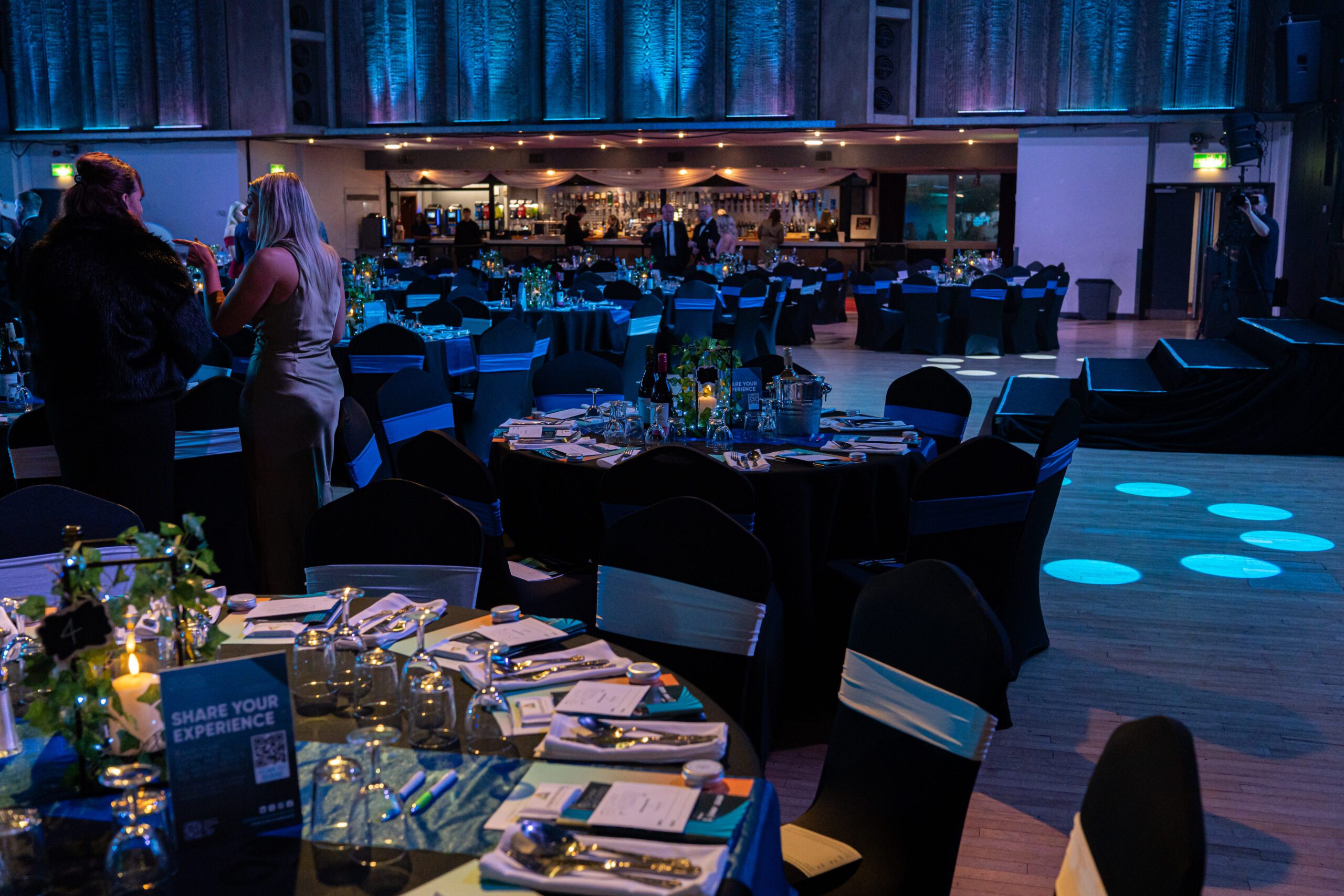 A dimly lit event venue with round tables set for dinner, black chairs with white sashes, and guests mingling near a bar in the background under blue lighting. A dimly lit event venue with round tables set for dinner, black chairs with white sashes, and guests mingling near a bar in the background under blue lighting.