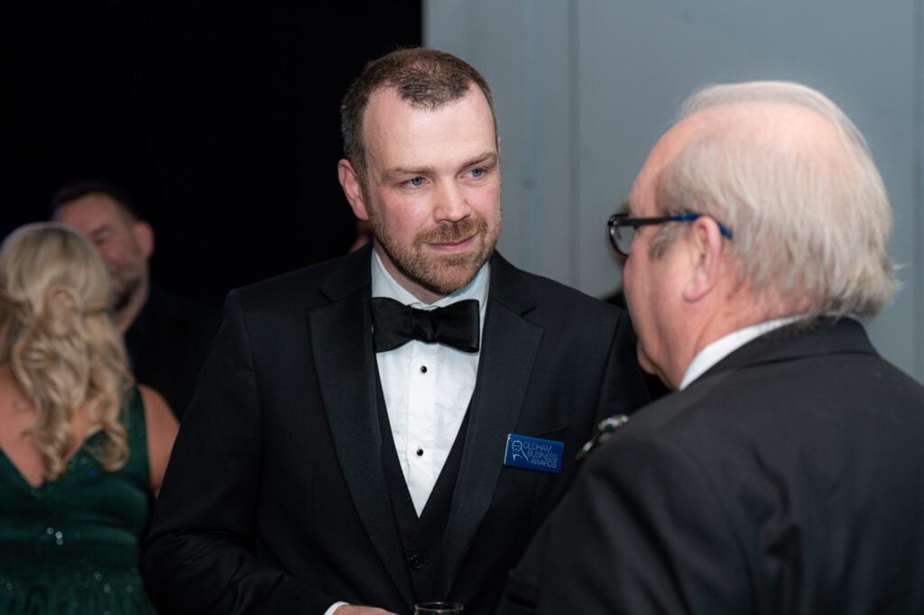 A man in a black tuxedo and bow tie talks to an older man in glasses at a formal event. Other people in evening attire are visible in the background. A man in a black tuxedo and bow tie talks to an older man in glasses at a formal event. Other people in evening attire are visible in the background.