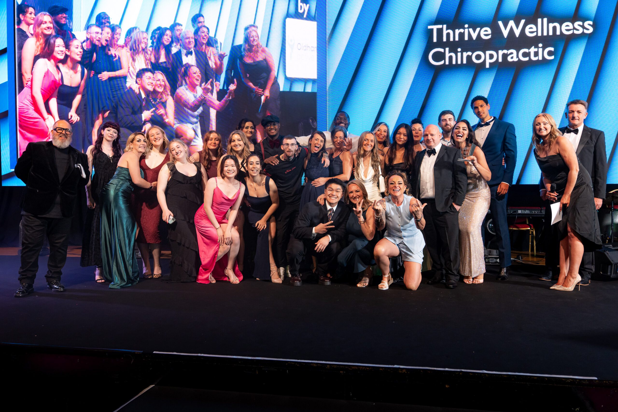A large group of people in formal attire pose together onstage, smiling and celebrating in front of a screen that reads Thrive Wellness Chiropractic.