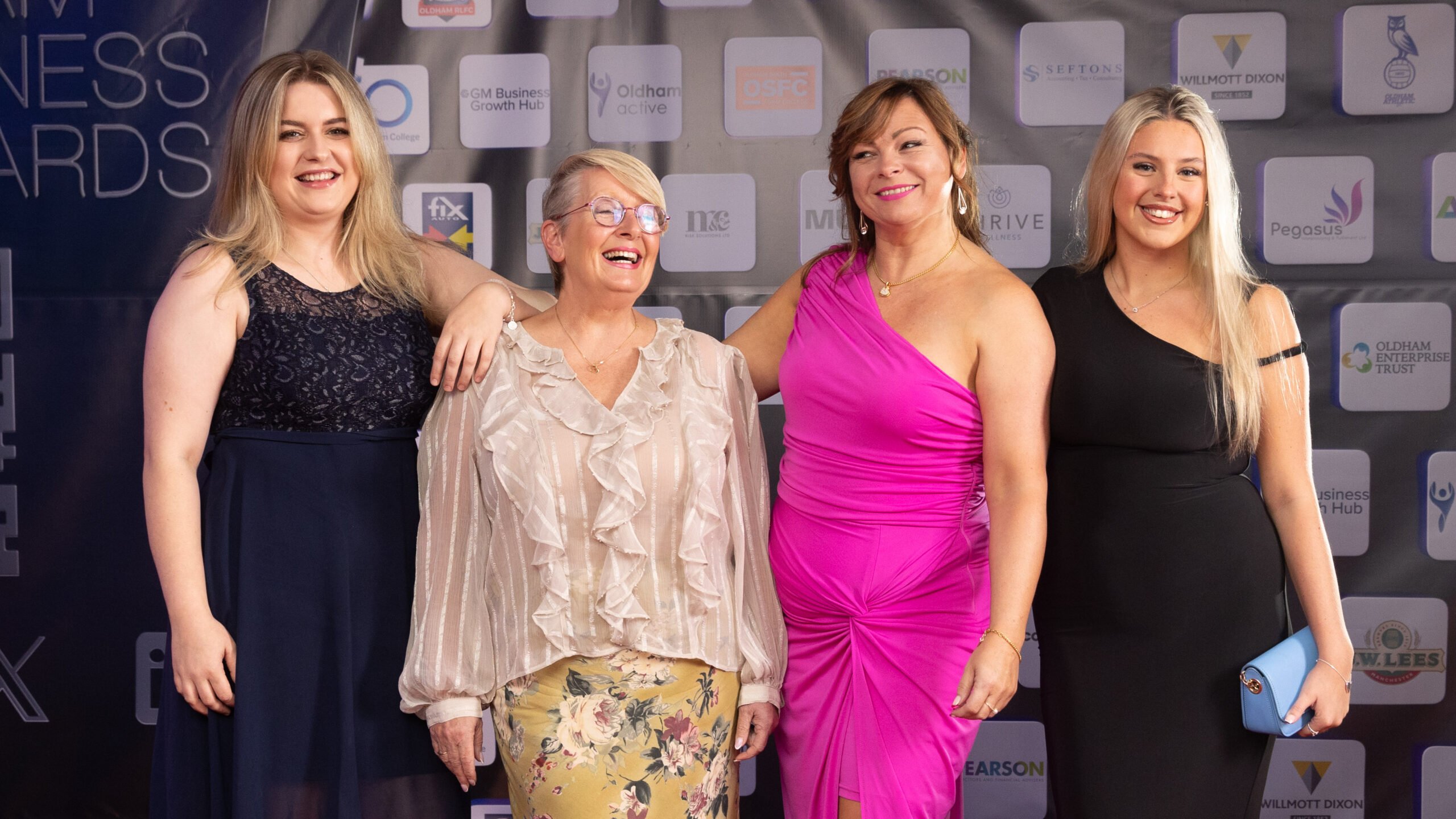 Four women pose together, smiling, at an event. They stand in front of a branded backdrop. Three wear dresses—one in navy, one in bright pink, and one in black—while the fourth wears a light blouse and floral skirt.