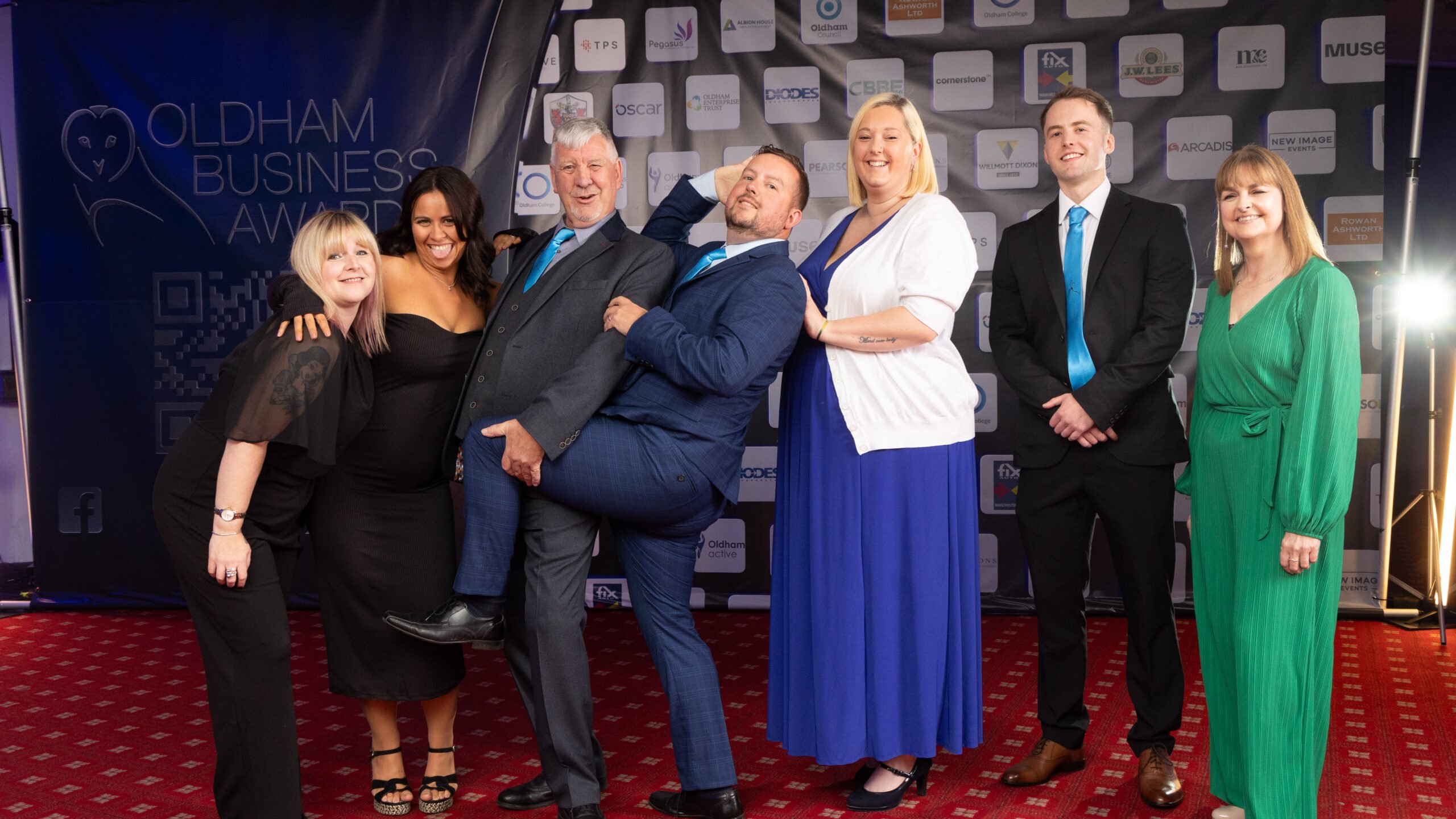 Seven people dressed formally pose and smile together at the Oldham Business Awards, standing in front of a backdrop with sponsor logos on a red carpet. One man playfully lifts his leg while others laugh.