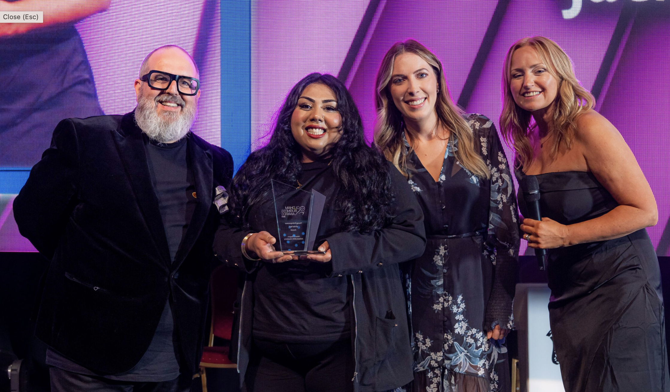 Four people stand together on stage, smiling at the camera. One person in the center holds a glass award. The background features colorful lights and a large display screen.